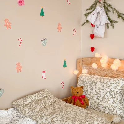 Wanddekoration mit Weihnachtsmotiv über einem Bett, mit Tannenzweigen und Lichtern som hänger darüber.