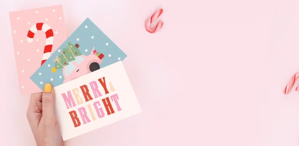 Hand, die bunte Geburtstagskarten mit Weihnachtsmotiven und den Texten „Merry Bright“ und „Ho Ho Ho“ auf einem Weihnachtsgeschenk hält.