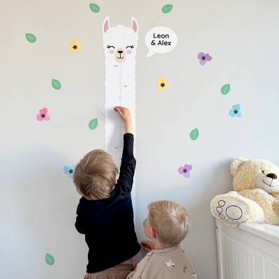Zwei Kinder messen ihre Größe an einer Wanddekoration in Form eines niedlichen Tiermotivs mit Namen, umgeben von bunten Blüten und Blättern, im Kinderzimmer. Perfekte Dekoration für spielerisches Lernen und persönliche Gestaltung.