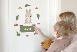 Türaufkleber mit Namen und einer süßen Kaninchenillustration für das Kinderzimmer, die eine Mutter zeigt, die auf die Dekoration zeigt, während sie ihr Kind und einen Teddybären hält. Perfekt zur Personalisierung des Zimmers für Kleinkinder.