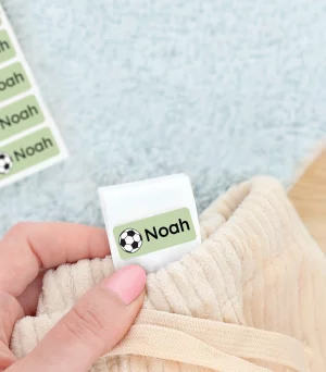 Personalisierte Namensetiketten für Kinder werden in diesem Bild angezeigt, während eines Sneakers auf einem weichen Teppich. Die Etiketten sind grün mit einem Fußballmotiv und dem Namen Noah darauf, ideal um Kleidung oder Schuhe zu kennzeichnen.