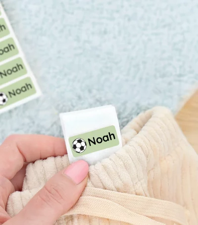 Personalisierte Namensetiketten für Kinder werden in diesem Bild angezeigt, während eines Sneakers auf einem weichen Teppich. Die Etiketten sind grün mit einem Fußballmotiv und dem Namen Noah darauf, ideal um Kleidung oder Schuhe zu kennzeichnen.
