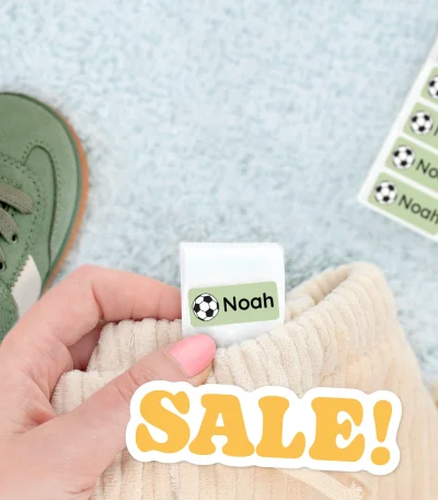 Das Bild zeigt eine Hand, die ein personalisiertes Namensetikett mit dem Namen Noah hält, das auf einem weichen Kleidungsstück angebracht ist. Im Hintergrund sind ein Paar grüne Kinder-Sneaker und weitere Namensetiketten sichtbar. Besonderer Fokus liegt auf den stilvollen und praktischen Namenslappen, ideal für die Kennzeichnung von Kinderkleidung und -spielzeug.