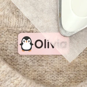 Personalisierte Namensetiketten mit einem niedlichen Pinguin-Design für Kinder. Ideal zum Beschriften von Kleidung, Spielsachen und mehr. Das Bild zeigt das Etikett auf einem weichen, strukturierten Untergrund.