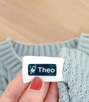 Individuelle Namensetiketten für Kinderbekleidung, die in einer Hand gehalten werden. Im Hintergrund sind herbstliche Blätter und ein weicher, blauer Pullover sichtbar. Ideal für die Personalisierung von Kleidung und Accessoires. Perfekt für Eltern, die ihre Kinderartikel kennzeichnen möchten.